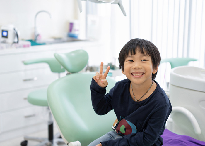 A joyful young boy in a dental clinic, smiling widely with a few missing front teeth, making a peace sign with his hand, sitting in a dental chair, with dental equipment in the background, conveying a positive pediatric dental visit.