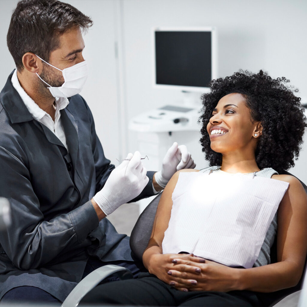 A cheerful young woman with curly hair smiles during a dental consultation with a male dentist in a modern clinic, both wearing masks, creating a friendly and reassuring atmosphere