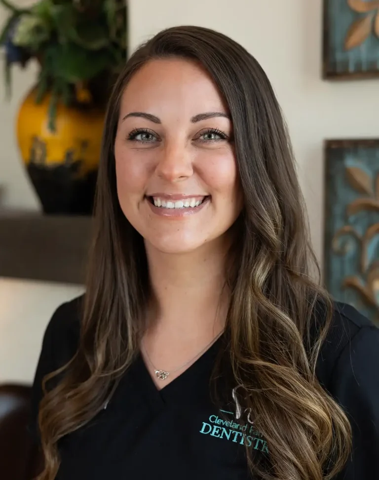 Shana smiles in a black branded top, presenting a warm and approachable presence in a dental office setting.