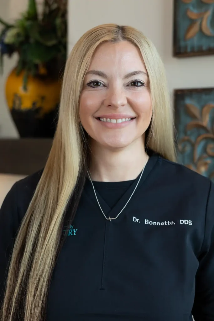 Dr. Carly Bonette smiles in a black branded top, presenting a friendly and professional appearance in a dental office setting.